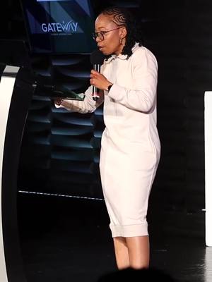 A woman in a white dress stands on a stage with a microphone, looking at a tablet she is holding. The stage has a dark background and a screen behind her with the word Gateway written on it. The lighting is focused on her face.