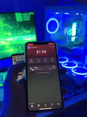 An individual holding a smartphone displaying a trading app interface. The background includes a computer monitor showing a cityscape, and a glowing computer case. The scene is bathed in a blue and green light.