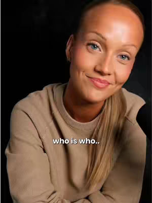 A woman with blonde hair, pulled back, is smiling gently, looking towards the camera. She is wearing a light brown sweater. A text overlay reading 'who is who..' appears near the bottom of the frame, and a dark backdrop fills the background.