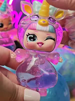 The cover image features a close-up of an LOL Surprise doll styled as a unicorn, held in a hand. The doll has a large, expressive face with big eyes, winking to the right. It is housed inside a transparent, pinkish-purple casing, giving it a liquid effect and a unicorn horn on top. The background is a blurred mix of purple, pink, and blue hues, suggesting a vibrant and playful setting.