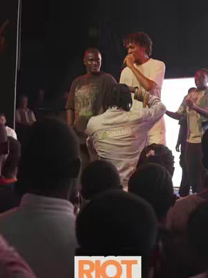 The cover image captures a live event with a clear focus on the stage and audience. The lighting is dim, directing attention to the performers on the stage. The performers include males, one holding a microphone and another wearing a T-shirt, which suggests casual attire. The audience is partially visible, creating depth and a sense of event.