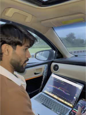 A man is seated in the driver's seat of a car, looking intently at a laptop displaying a financial chart in the foreground. A smartphone is also visible in the car. The interior of the car and the passing scenery outside are visible.