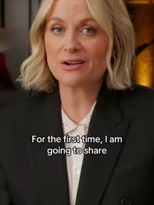 A medium shot of a woman speaking, with text overlayed at the bottom. She has blonde hair, is wearing a black blazer over a white collared shirt and has gold hoop earrings. The background appears to be a neutral, warm-toned setting.