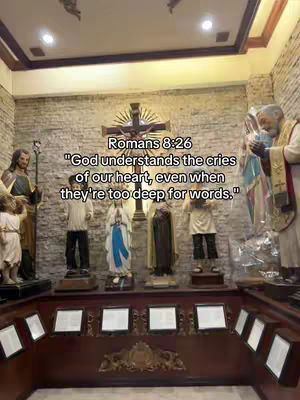 An interior shot inside a church or religious building, featuring various statues and religious figures. The composition is centered, with the statues lined up on a raised platform, and a large cross in the background. The color palette is muted, with warm tones from the wooden elements. Text is overlaid, displaying a Bible verse.
