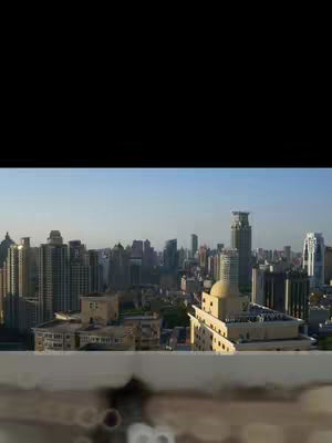 An eye-level shot of a densely packed cityscape under a clear, light-blue sky. Numerous high-rise buildings dominate the frame, varying in height and design, with an array of windows reflecting the daylight. The composition includes a low foreground element, possibly a rooftop, and bokeh lights.