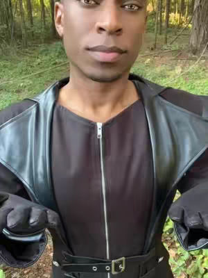 A medium shot of a Black man in a black outfit with a leather-like vest in front of a forest backdrop. The man is looking towards the camera. This appears to be a selfie video.