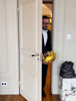 A man in a tuxedo and a white bow tie opens a white door. He holds a gold balloon that says Happy New Year. Behind him, you can see an interior hallway with a doorframe and some lights. There is also an off white wall wainscotting. To the right of the door, on a white marble pedestal, is a coiled stack of black cables.