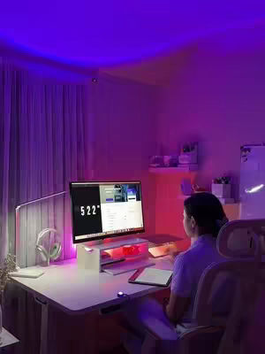 A woman sits at a desk in a room lit with purple and pink lights, focused on a computer screen. Her workspace includes a monitor stand with what appears to be a tablet beneath it, a lamp, and a pair of headphones. The scene has a modern, study-focused aesthetic.
