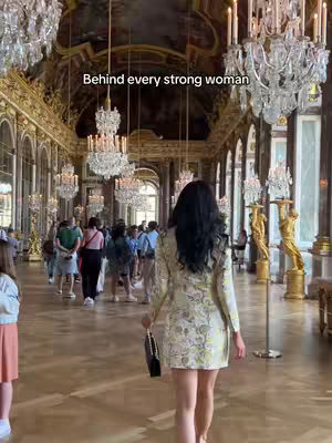 A person with long dark hair, wearing a patterned dress, walks away from the camera down a grand, ornately decorated hallway. The hallway is lined with people, chandeliers, and classical sculptures. The setting suggests a historical or palatial interior.