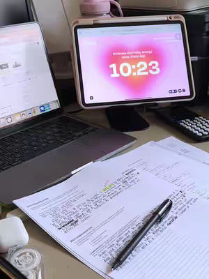 The image shows a desk setup with a laptop, a tablet displaying a heart-shaped timer, and some papers with notes on them. A pen and a calculator are also visible. The color scheme is neutral, with touches of pink and white.
