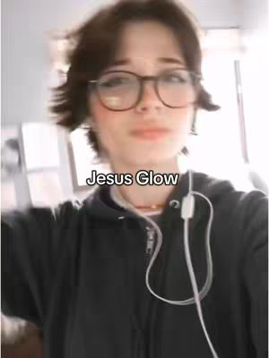 A slightly blurry selfie of a young person with short, dark hair, wearing glasses and a black hoodie. The text overlayed says Jesus Glow.