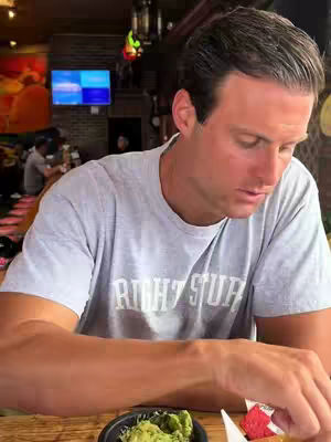 A man sits at a table in a restaurant, looking down. He wears a grey shirt; the restaurant has a TV showing content.
