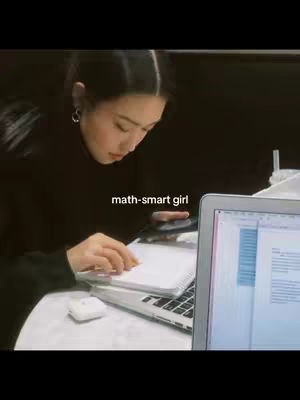 A young woman is seated at a table, appearing to be studying. She is leaning over a notebook, with a laptop open on the table in front of her, displaying what looks like a document. The lighting suggests a dimly lit setting, possibly a cafe or study area, giving a focused, studious atmosphere.