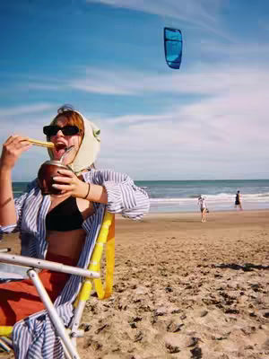 A vibrant beach scene with a woman enjoying a drink, a kite in the sky and two other people at the background. The woman, dressed in a shirt and shades, is seated in a beach chair, holding a vessel with a straw to her mouth. The sky is a bright blue, the ocean and beach sands fill the background.