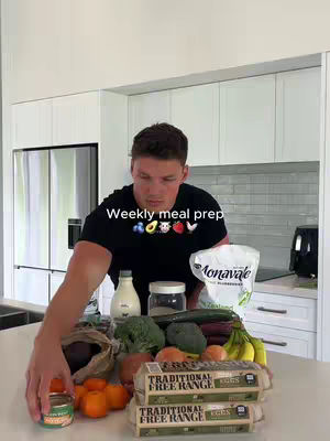 A man in a black t-shirt stands in a kitchen with various food items on a countertop, including fruits, vegetables, eggs, and packaged goods. The background includes white cabinets, a stainless steel refrigerator, and a tiled backsplash. Text overlay describes this as a weekly meal prep.