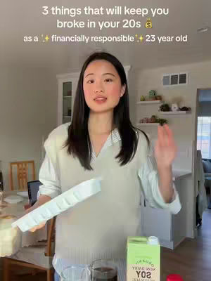 A woman is standing indoors, facing the camera with a neutral expression, left arm slightly raised and a hand gesturing. She is wearing a light-colored sweater over a collared shirt, with long dark hair. In the background, a kitchen setting is visible, and text overlays at the top.