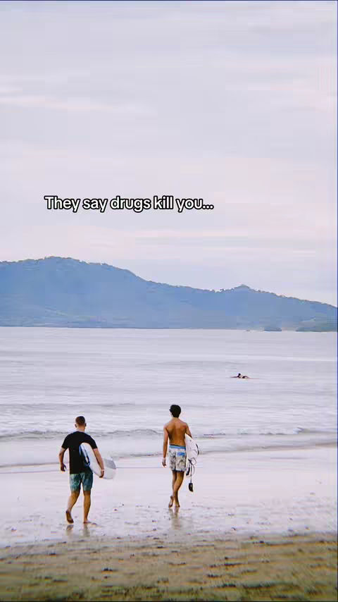 Two men walk on a beach carrying surfboards. Text overlayed at the top reads, "They say drugs kill you...". The sky is overcast and the water is a soft grey.