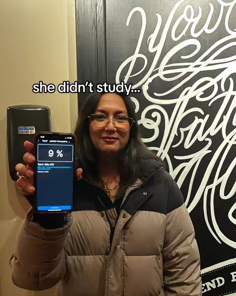 A woman stands in front of a graffiti-covered wall, holding a smartphone displaying a grade of 9%. Text overlay 'she didn't study...' is at the top. She is wearing glasses and a puffer jacket.