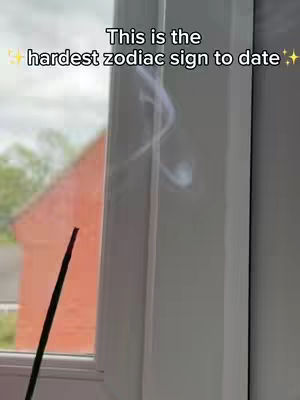 The image features a window with white curtains, behind which a red building is visible. A black stick is positioned diagonally in the left foreground. The text overlay indicates the video's focus on zodiac signs.
