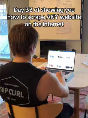 A person is sitting at a table, facing away from the camera, working on a laptop. The text overlay reads: Day 33 of showing you how to scrape ANY website on the internet. The person wears a black sleeveless shirt. The laptop screen shows a website with multiple sunglass models. The setting appears to be a home office or study, with a whiteboard and window visible in the background.