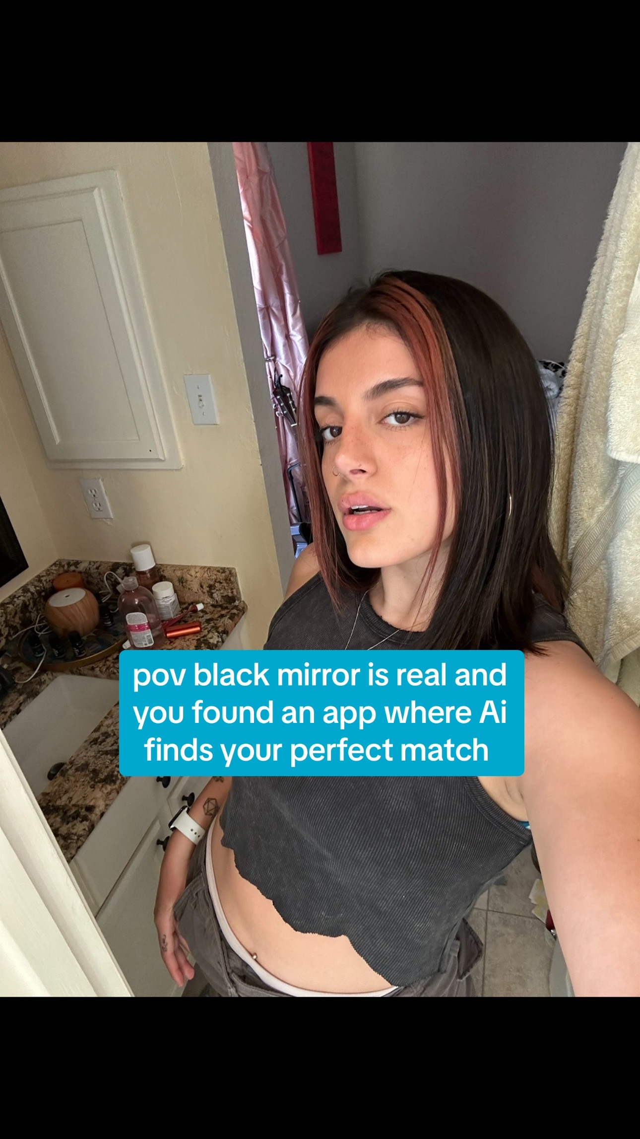 A selfie-style image featuring a young woman in a bathroom setting. The woman, with dark hair and red-toned highlights, looks directly at the camera with a slight, neutral expression. She wears a dark, ribbed tank top, and her lower torso is visible, with a tattoo on her left arm. The turquoise text overlay states, "pov black mirror is real and you found an app where AI finds your perfect match."