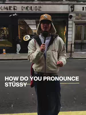 A man standing in front of a building with text overlayed that asks how to pronounce the brand name Stussy. He is wearing a Yankee baseball cap, a beige jacket, a white shirt, and a long dark-colored skirt or bottom. He is holding a microphone. The overall composition is a selfie with a background.