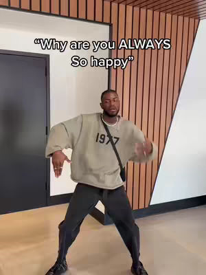 A man in a casual outfit is dancing energetically in front of a neutral background with a wooden panel wall to the right. The text overlay says, “Why are you ALWAYS so happy”.