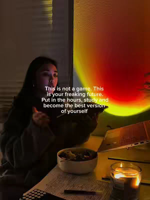 A person, lit by warm light, sits at a desk with a laptop and study materials, appearing to be engrossed in focused study. A warm-colored light, possibly from a sun lamp, casts a glow on the wall. Text overlayed on the image features motivational messages.