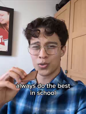 A young man with glasses holds a piece of food, likely a cookie, and looks at the camera. Text overlayed on the bottom says "always do the best in school". He is in a room with a framed picture and wooden cabinets behind him.