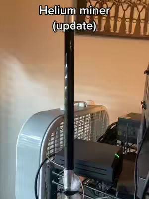 The cover image shows a close-up of a Helium miner setup. A black cylindrical pole is the central element, likely an antenna, with a light blue fan and other electronic components behind it. The scene is set indoors with a beige wall and a framed artwork in the upper right corner.
