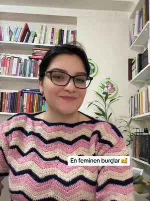 A woman with glasses smiles at the camera. She is wearing a pink and white patterned sweater. Behind her are bookshelves filled with books.