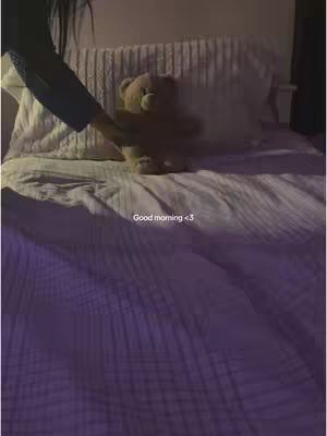 The image shows a bedroom scene in soft light. In the foreground is a bed covered in white sheets with a teddy bear sitting upright on top. A hand with a dark sleeve is gently touching the bear. The text 'Good morning <3' is added below the bear and is softly visible.