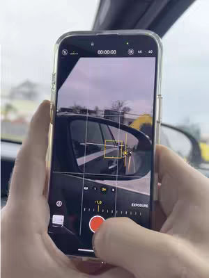 A person holds a phone with the camera app open, recording inside a car. The phone screen displays a view through the car's mirror. The image is overall cool-toned, with the focus on the phone and the car interior.