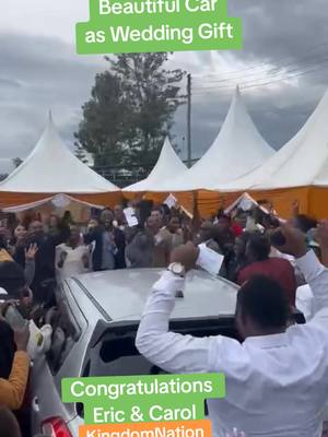 A crowd celebrates at an outdoor event, likely a wedding. A silver car is surrounded by people, with several white tents in the background. Text overlays indicate that the car is a wedding gift and congratulates the couple, Eric and Carol, with the hashtag #KingdomNation.