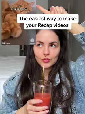 A selfie-style image featuring a woman holding a glass with a red liquid and a gold straw. She is looking slightly to the side, with a small image in the top left corner that seems to be a screen recording of a cat in a background with text that reads 'My 2023 so far'. The background includes a blurred interior with white bedding or upholstery, adding a soft contrast to the bright colors of the drink and the woman's denim jacket.
