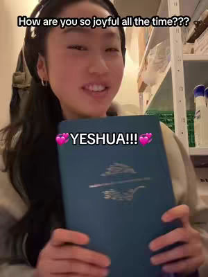 A young woman with long black hair and a headset is smiling and holding a blue book with the word YESHUA on it and two hearts superimposed on the photo. The background appears to be a room with shelves and other items.
