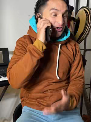 A man in a brown hoodie with a light blue inner layer, holding a phone to his ear. He is looking towards the camera with his mouth slightly open as if he's talking.