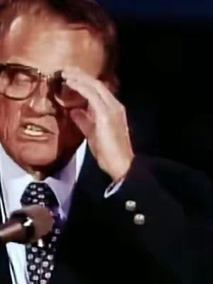 A medium shot showing a man with glasses, wearing a dark suit with a patterned tie, touching his forehead with his right hand, and speaking into a microphone. The background is dark.
