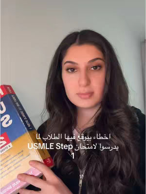 A young woman with long, dark hair is centered in the frame, holding a textbook. The background is a plain, light-colored wall. On the cover of the book, there's a red and blue design with some text visible. The woman's expression is neutral, and she's looking directly at the camera. The image is well-lit, and the composition focuses on the woman and the book she's holding.