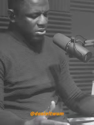 A black and white image featuring a man speaking in front of a microphone. He has short hair and is wearing a textured dark-colored sweater. The microphone is positioned near his mouth, suggesting he's either singing or speaking into it.