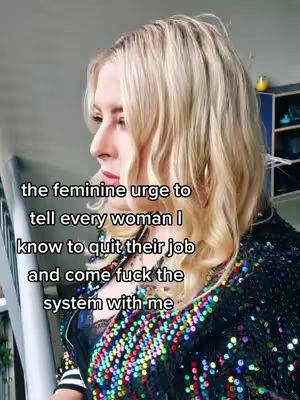 A woman with blonde hair faces the left, looking slightly off-camera. She wears a dark, sequined top with a colorful neckline and a black top underneath. The background is blurred with a gray surface and a glimpse of a shelf with a yellow object.