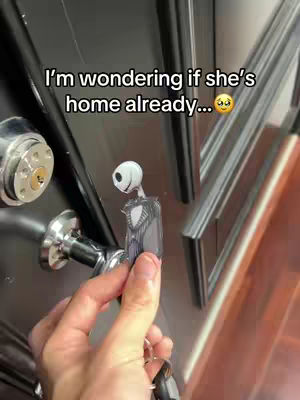 The image shows a hand holding a key adorned with a Jack Skellington-themed charm near a black door with silver fixtures. Text overlay states, "I'm wondering if she's home already...", suggesting a playful, anticipatory mood.