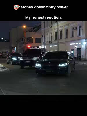 The image shows a street scene at night with several vehicles, primarily dark-colored cars, in the foreground. The street is illuminated with streetlights, and buildings line both sides. Text overlays the top, including an emoji and a message.