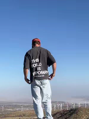A person stands with their back to the camera on a hill overlooking a landscape of wind turbines under a clear blue sky. They're wearing a dark t-shirt with white text and light-colored jeans, and a red cap. The scene is bathed in natural light.