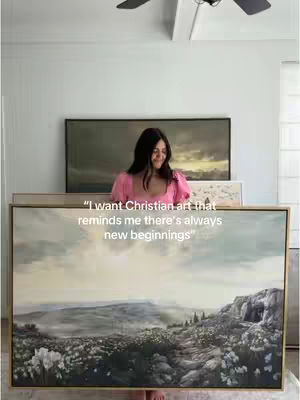 A woman stands in a brightly lit room, holding up two pieces of framed artwork. The woman is smiling, and behind her is a large black framed artwork. The artwork at the bottom is landscape with what looks like a mountain. The background is a light color with a ceiling fan.