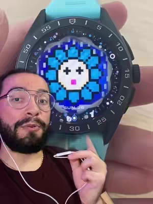 A man with glasses holds a smartwatch displaying a pixelated flower design on its screen, with the man's face and the watch prominent in the image.