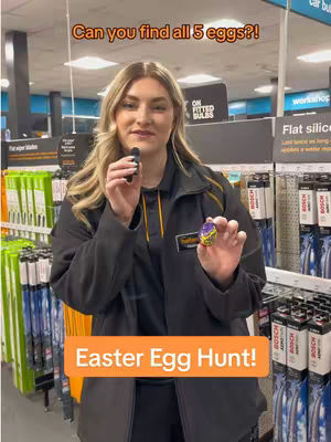 A woman with blonde hair wearing a black jacket holds a small, decorated Easter egg in front of a store display. Text overlay announces an "Easter Egg Hunt!"