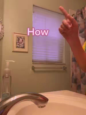 The image is a selfie-style shot taken in a bathroom. The main subject is a hand pointing upwards, with the index finger extended. The background includes a framed picture, a closed window with blinds, a towel rack, a sink, and a soap dispenser. Text overlayed on the image says 'How'. The overall color palette appears to be light and clean with a cool, slightly purple hue.