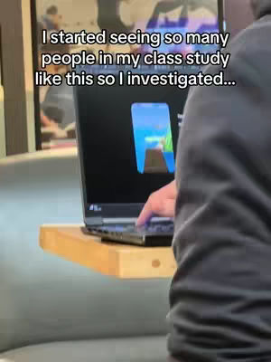 A person is shown from the chest up working on a laptop on a wooden surface. The person's right hand is on the laptop, and the screen displays a blue and white graphic. Behind the laptop, a section of a wall art can be partially seen. The image has subdued lighting with a slightly out-of-focus background.