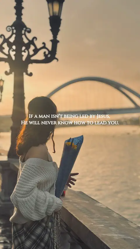 A woman stands on a stone ledge, looking away from the camera towards a body of water. She holds a bouquet-like arrangement. The background features an arching bridge and a sunset sky.